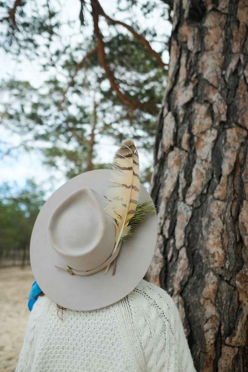 A woman in a feathered hat standing in front of a a tree.