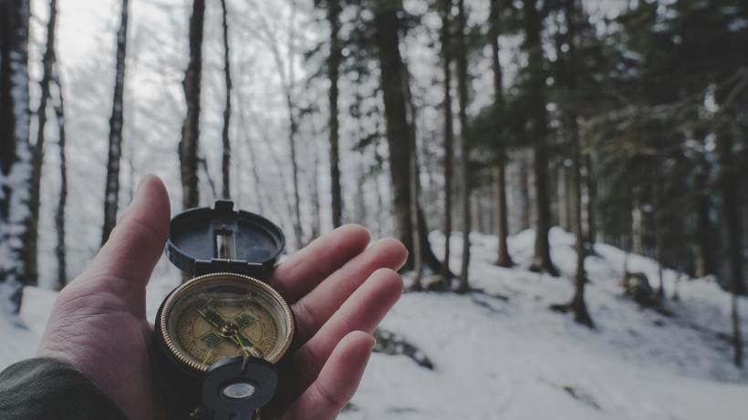A person's hand holding a compass in the middle of a snowy forest.